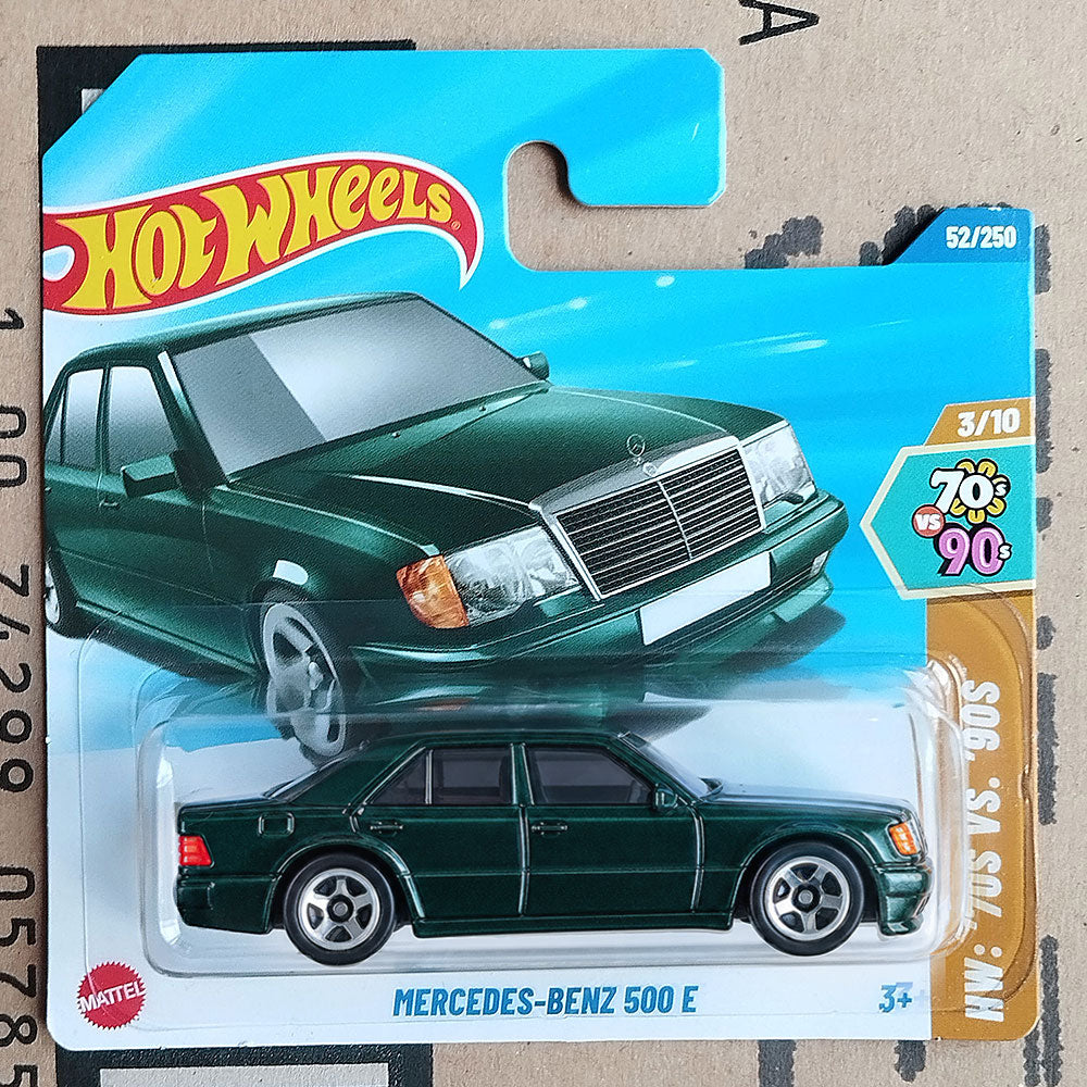A Hot Wheels die-cast toy car of a dark green Mercedes-Benz 500 E from the 1990s, presented in its original blister card packaging on a cardboard background.